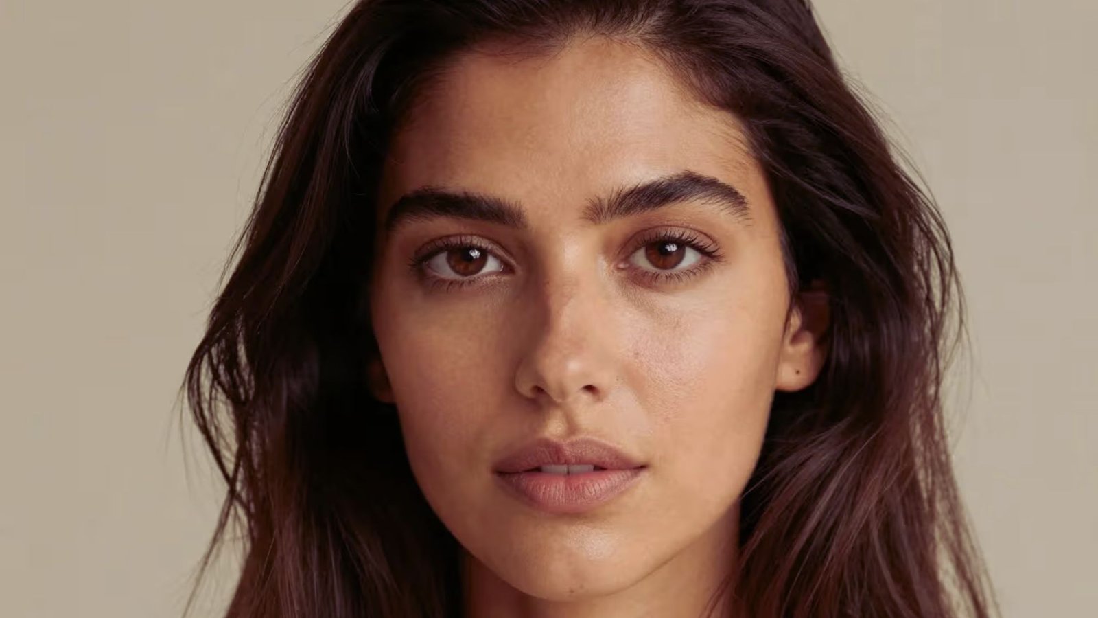 A woman with long brown hair wearing a white top, standing against a neutral background.