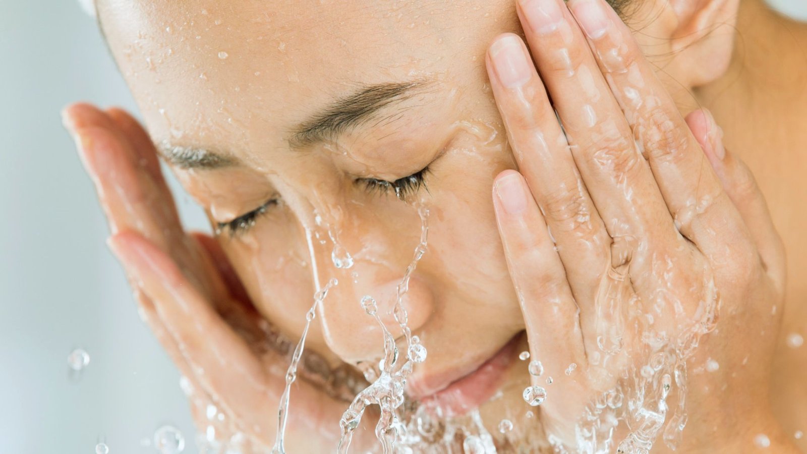 A woman is gently washing her face with water at a bathroom sink, focusing on her skincare routine.