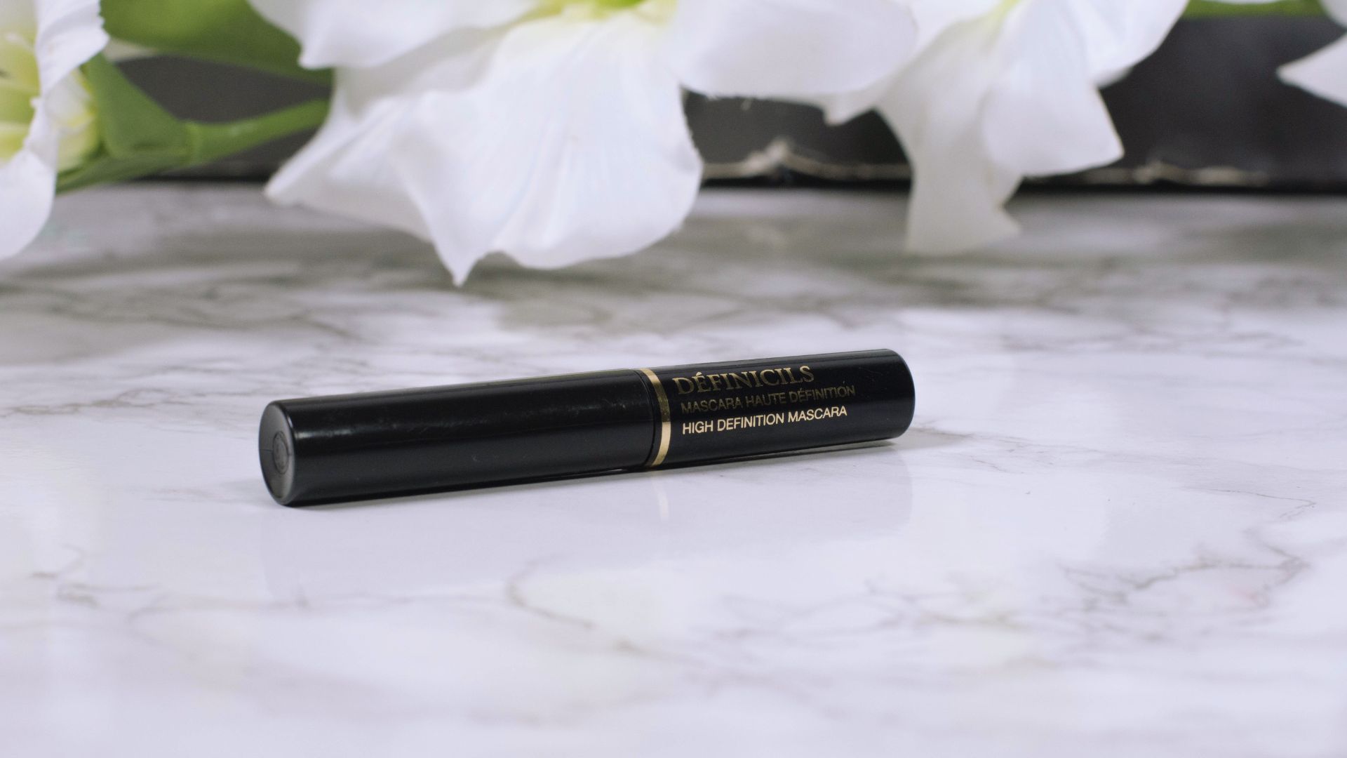 A lipstick tube rests on a marble counter, surrounded by delicate white flowers.