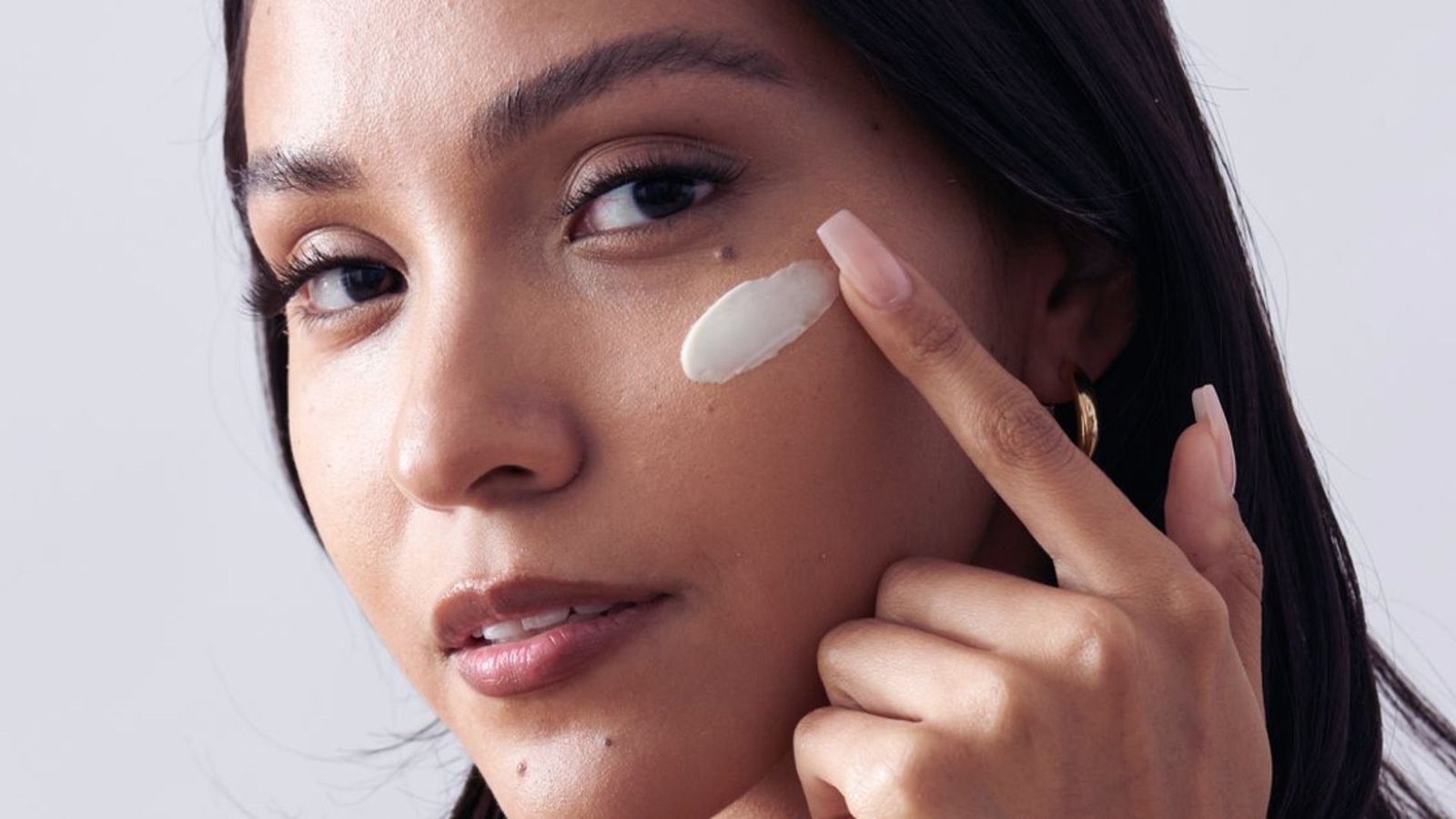 A woman gently applies cream to her face, focusing on skincare in a well-lit bathroom setting.