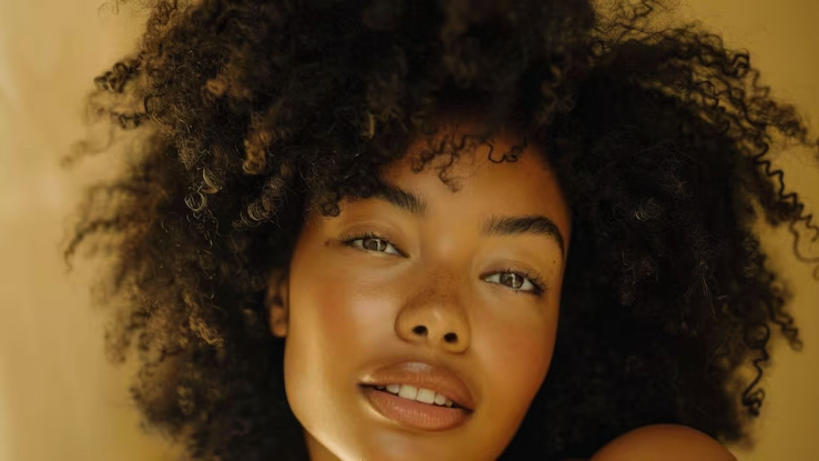 A beautiful woman with curly hair and a tan smiles warmly at the camera, showcasing her radiant complexion.