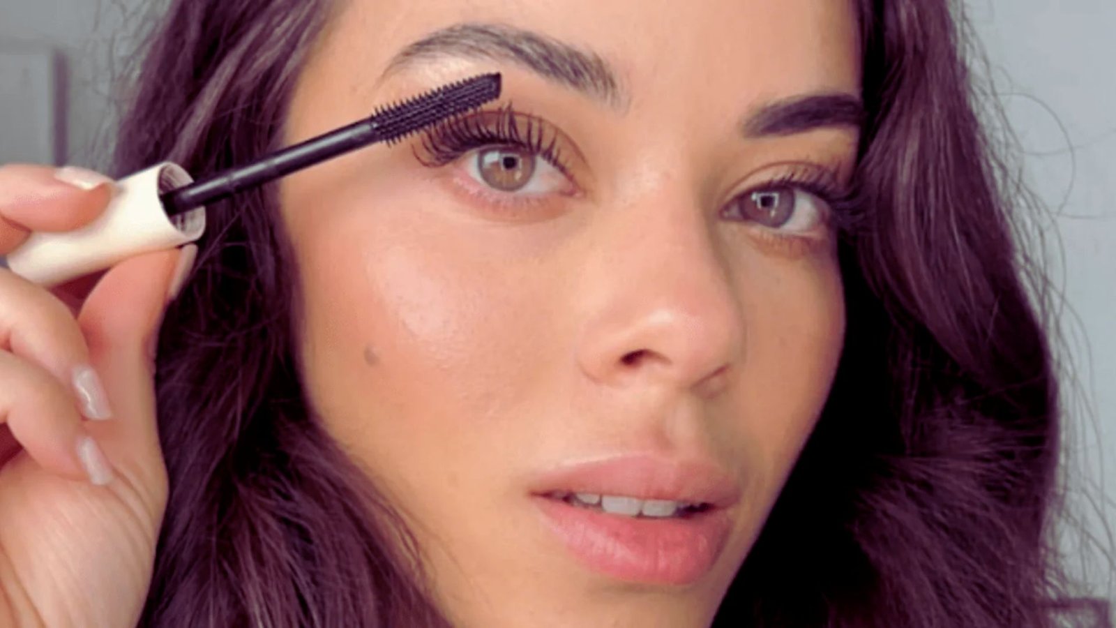A woman applies mascara to her eyelashes using a mascara brush, focusing intently on her makeup application.