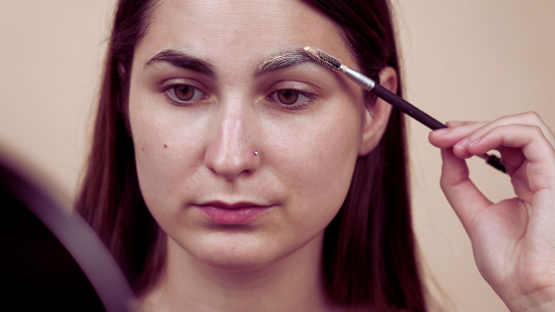 A woman carefully applies makeup to enhance her eyebrows in a well-lit setting.
