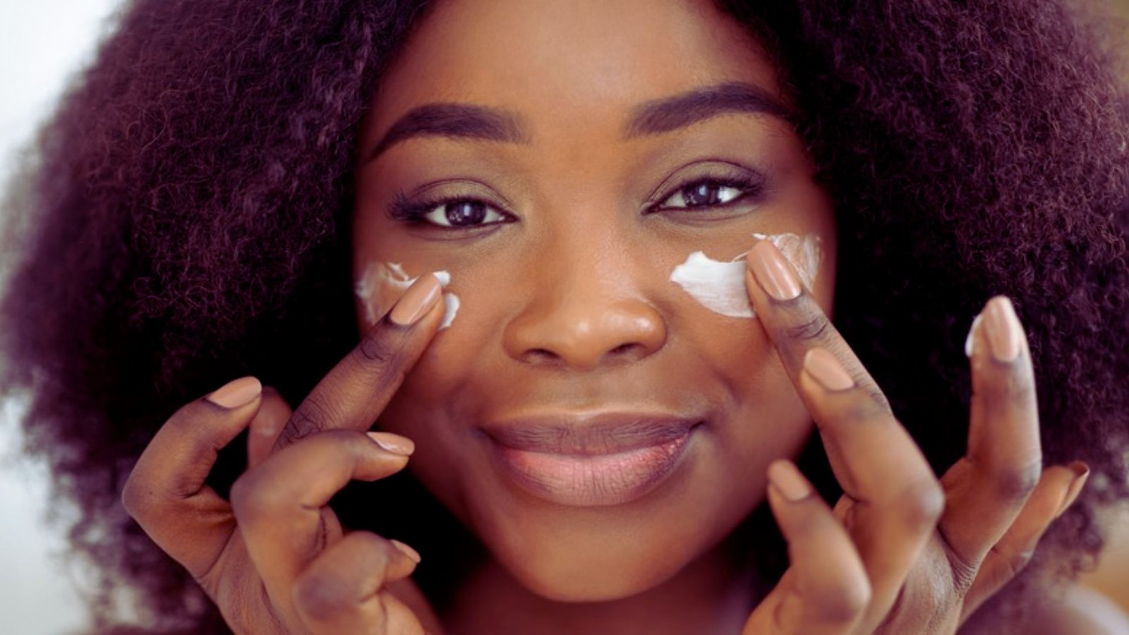 A woman with a black afro holds up a cream product, smiling and showcasing it to the camera.