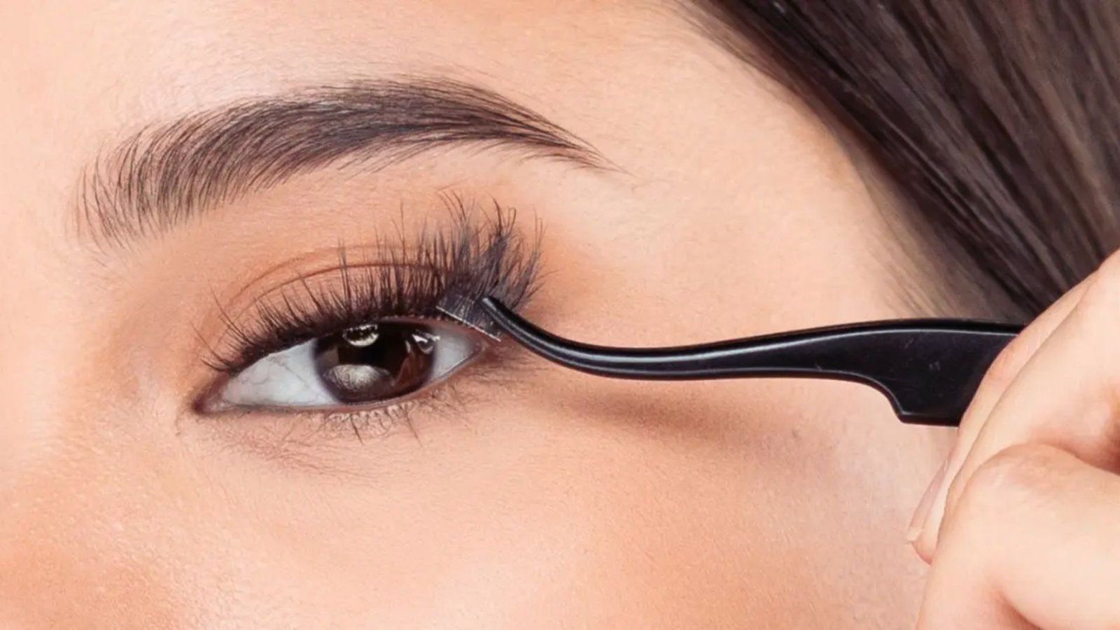 A woman applies mascara to her eyelashes using a brush, focusing intently on her eye in a well-lit setting.