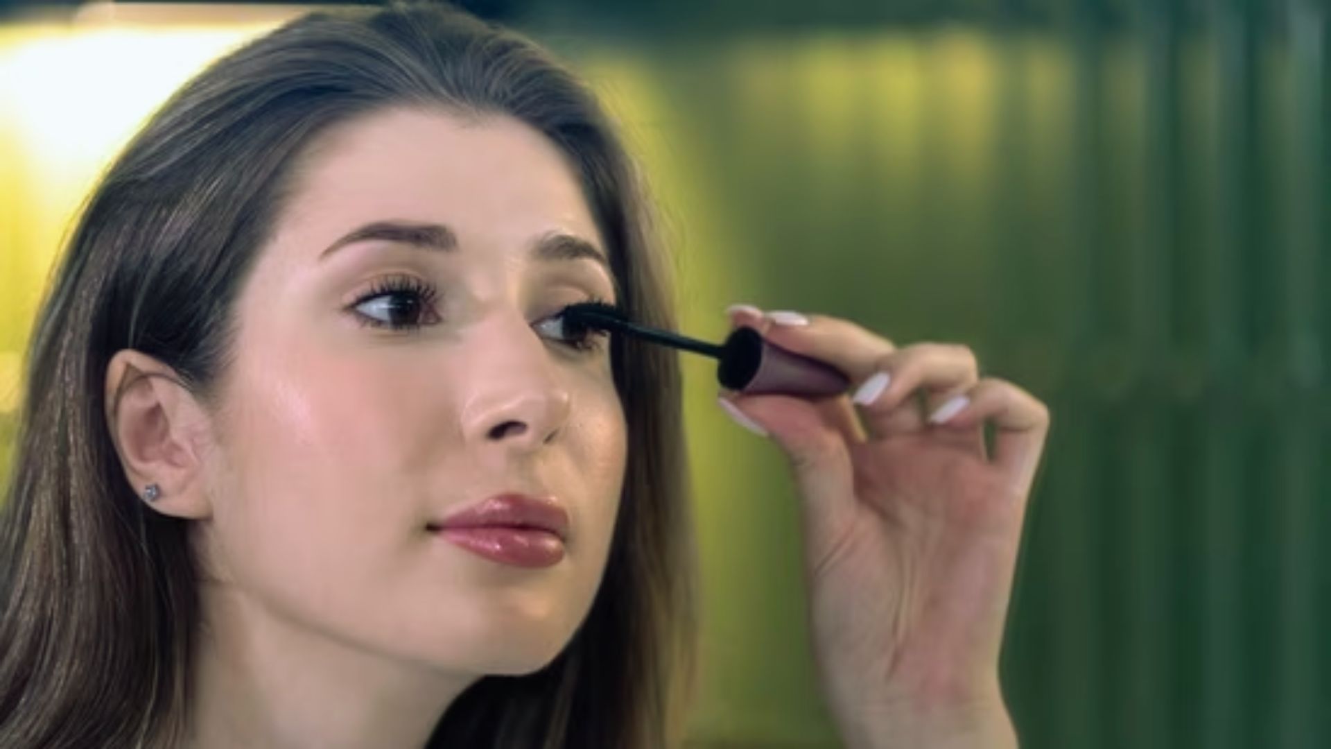 A woman carefully applies mascara to enhance her eyelashes in a well-lit bathroom mirror.