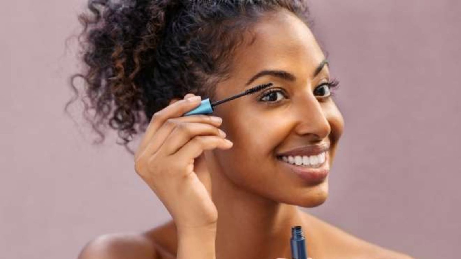 A woman smiles while applying mascara with a brush, enhancing her eyelashes in a bright, well-lit setting.
