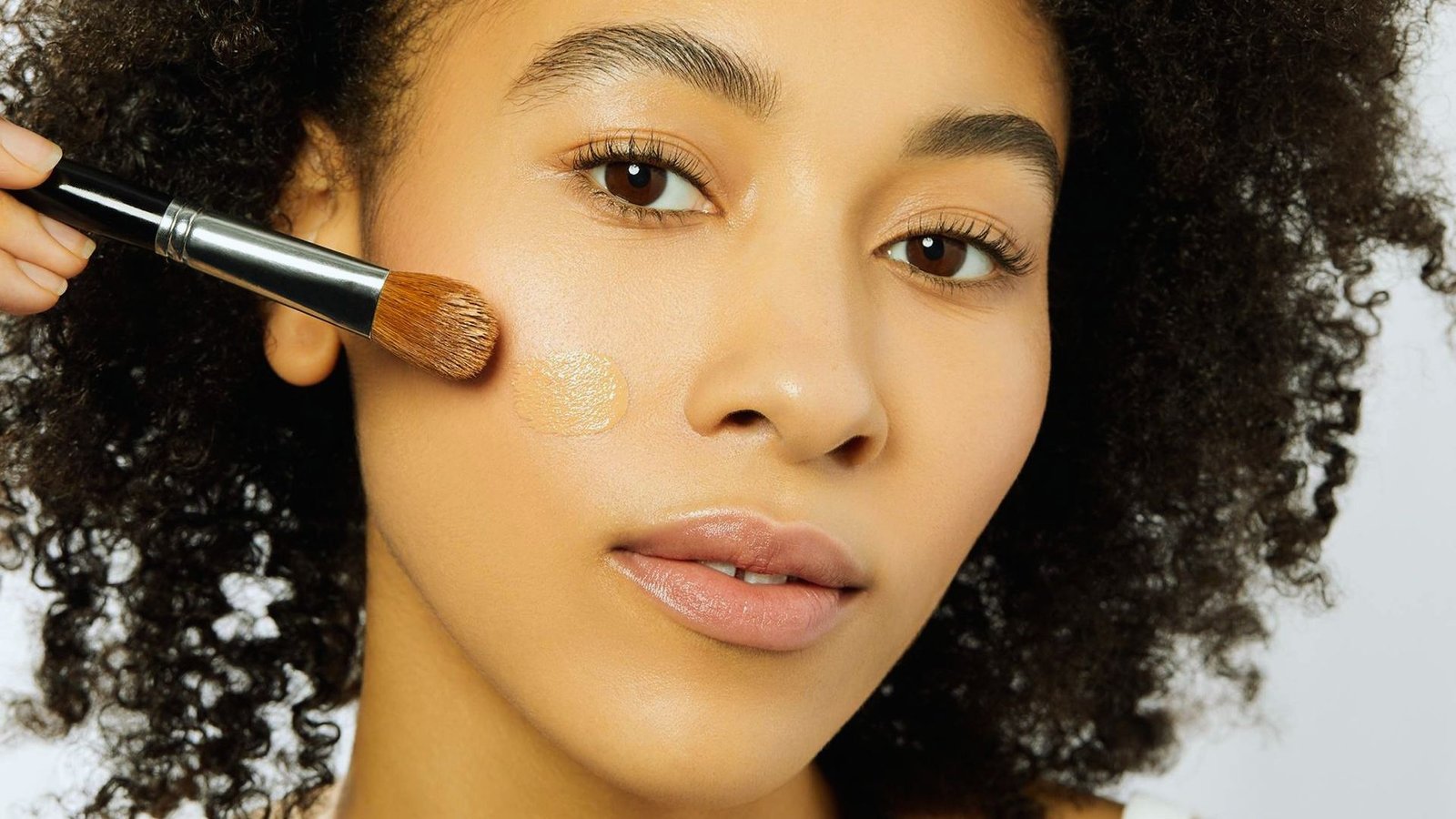 A woman with curly hair applies makeup in front of a mirror, focusing on her eyes with a brush.