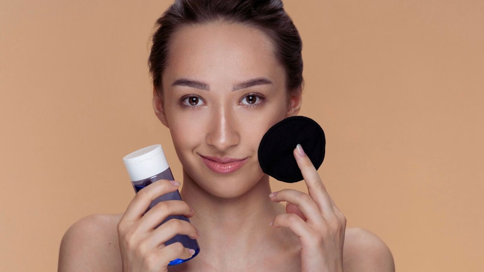 A woman holds a sponge in one hand and a bottle of face wash in the other, preparing for her skincare routine.