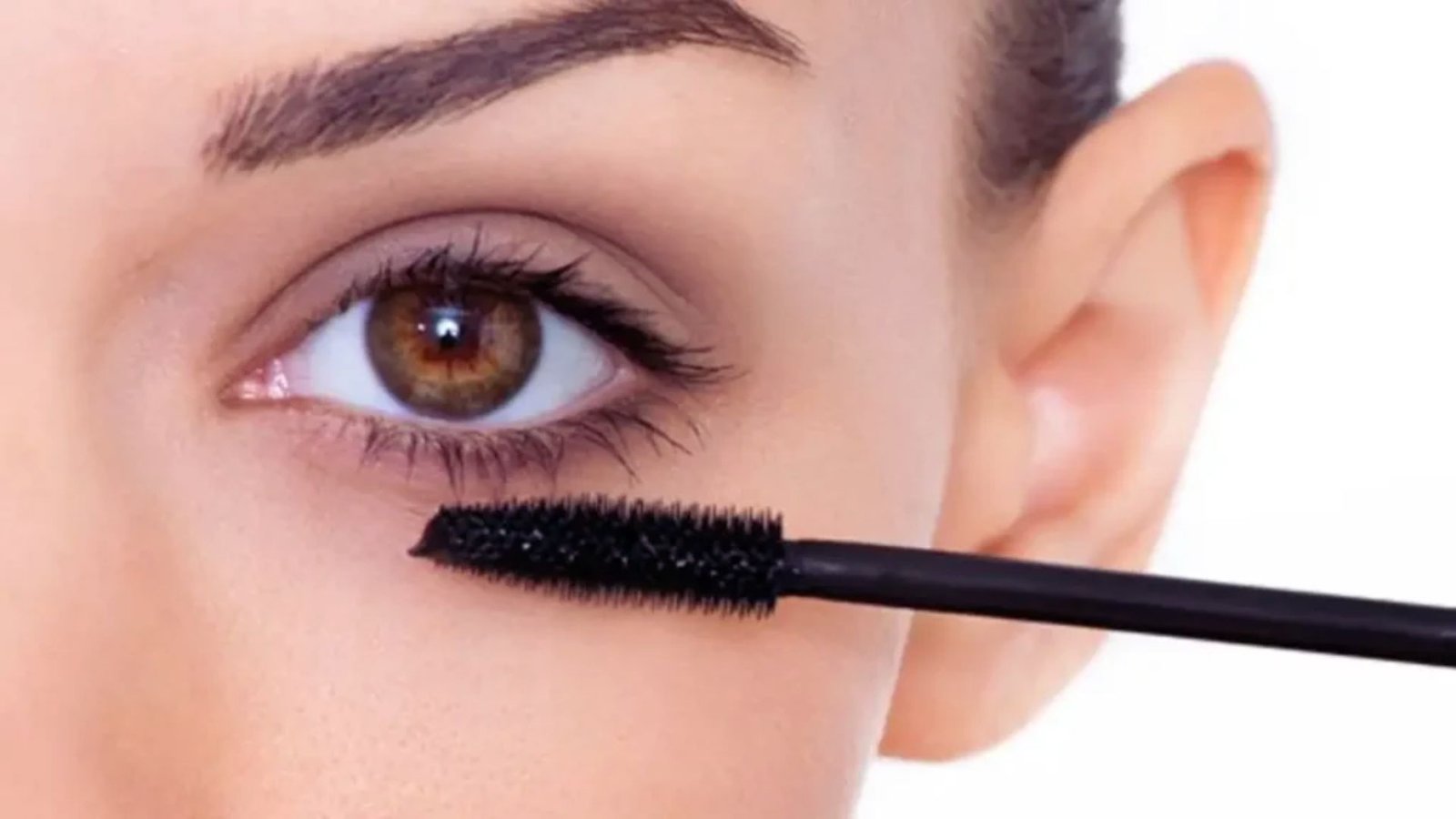A woman applies mascara to her eyelashes using a mascara brush, focusing intently on her makeup application.