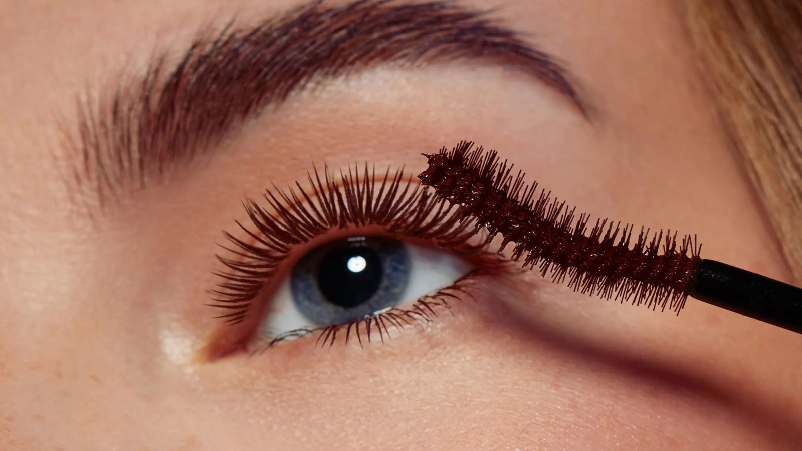 A woman applies mascara to her eyelashes, enhancing her eye makeup in a well-lit bathroom setting.