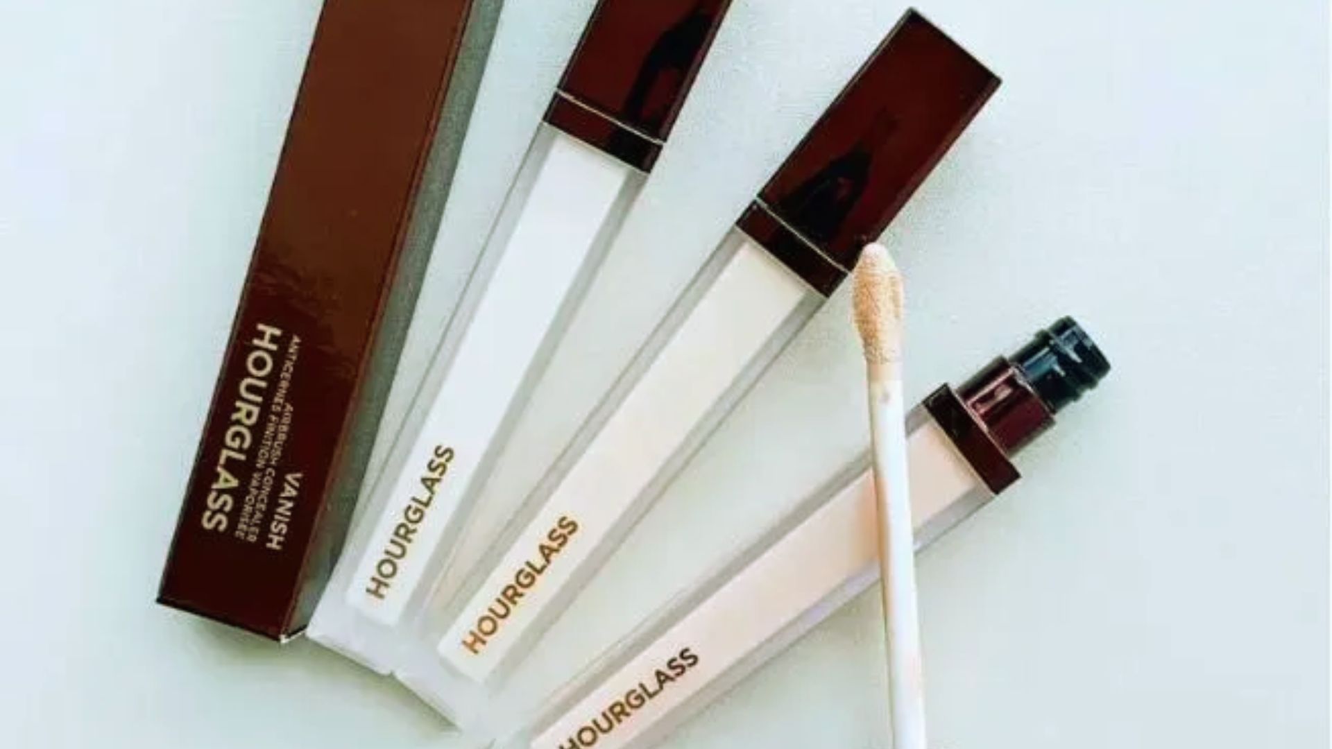 Three types of concealers in various shades alongside a makeup brush on a neutral background.